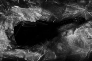 Photographie noir et blanc réalisée en Blind Painting d'une guitare probablement de dos. La guitare est très noire et semble posée sur un écrin blanc et vaporeux,qui n'est pas sans rappeler un cercueil ou quelque chose de funéraire.